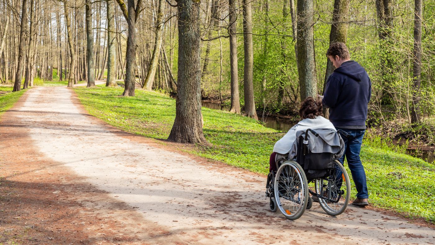 Två personer, en i rullstol promenerar på grusväg i vårgrön skog.