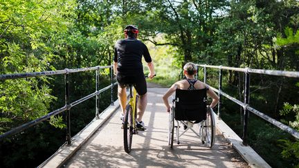 Två personer, på cykel och rullstol korsar en liten bro. 