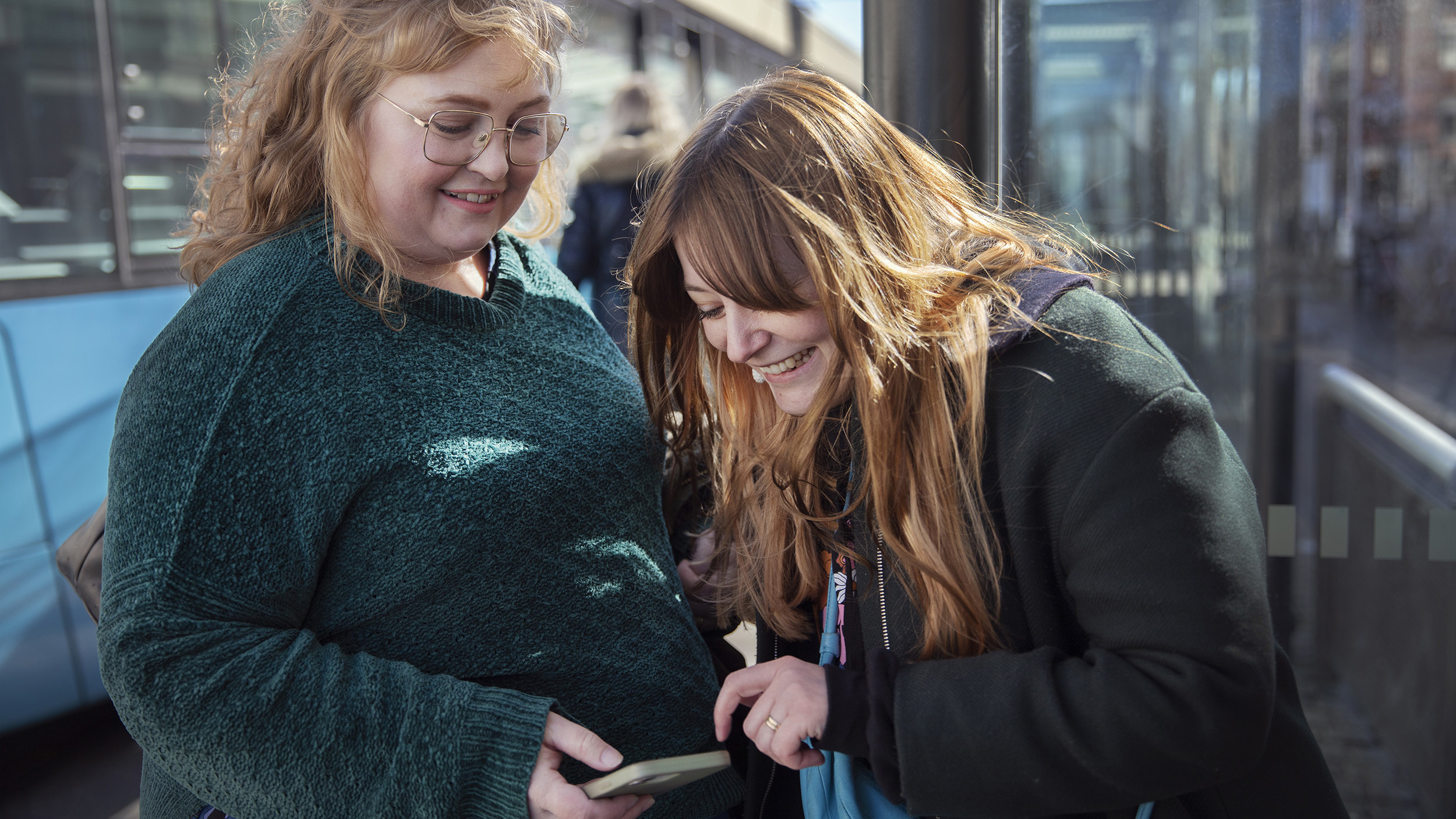 Två personer på en busshållplats tittar på en mobil och skrattar.