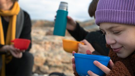 Ett barn som dricker ur en kåsa och ler, i bakgrunden detaljer av andra med kåsor och termos.