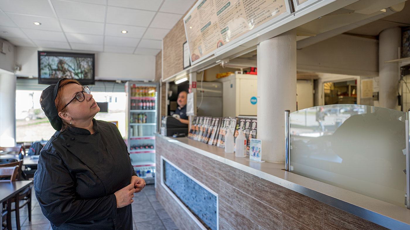 en kvinna i svarta arbetskläder står på en pizzeria och tittar upp mot tavlan med menyn.