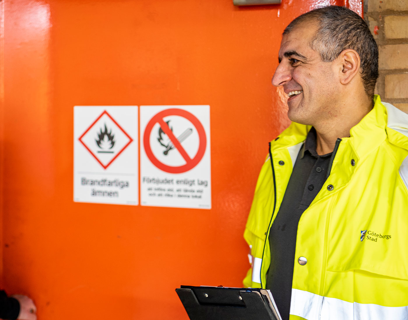 En man i gul signaljacka ler mot någon. I handen har han en skrivplatta. Han verkar befinna sig på någon sorts industri.