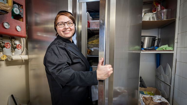 En glad kvinna i svarta kökskläder öppnar en kylskåpsdörr samtidigt som hon ler mot kameran.
