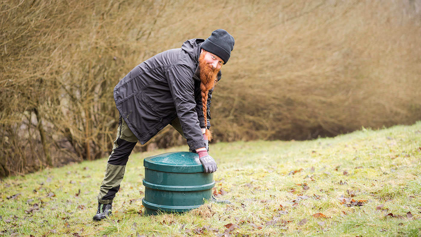 En leende man i oömma kläder och långt rött flätat skägg lyfter på ett brunnslock samtidigt som han tittar mot kameran.