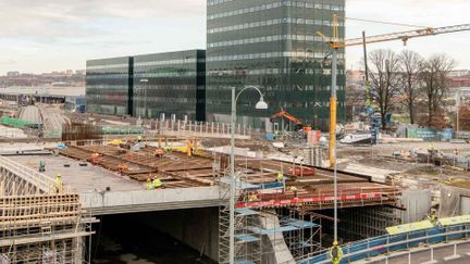 Byggarbetsplats med glasbyggnader i bakgrunden och en ofärdig konstruktion i förgrunden.
