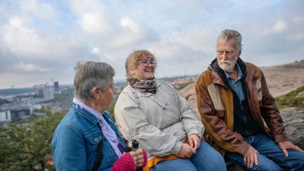 Ett foto av tre personer av olika generationer som sitter på en höjd. I bakgrunden syns grönska och byggnader.