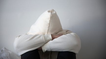 En person sitter med huvudet tungt lutat mot korslagda armar och uppdragna knän. Personen är klädd i vit hoodie och svarta byxor. Obestämt kön och ålder.
