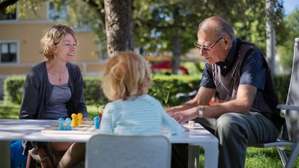 Tre personer av olika generationer som sitter runt ett bord i barnhöjd utomhus.