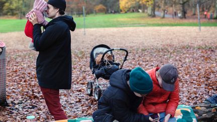 En vuxen håller upp ett barn i famnen i en park, medan två andra personer sitter på en bänk och tittar på en mobil.