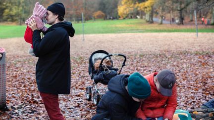 Familj i Slottskogen en höstdag. En pappa lyfter upp sitt barn ur en barnvagn äldre barn läser i förgrunden av bilden.