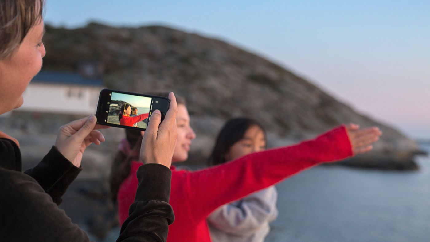 en flicka med rödtröja pekar med sin arm ut över havet. En kvinna i förgrunden av bilden tar ett kort på henne.