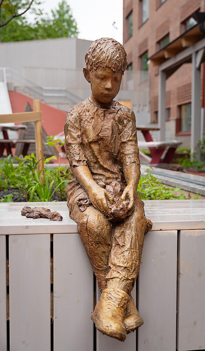 En staty av en pojke som sitter p&aring; en b&auml;nk.