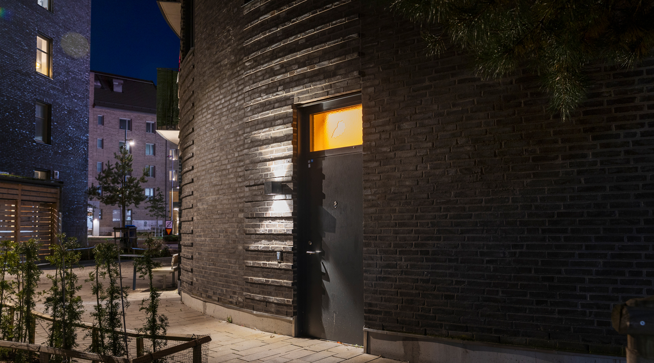 En mörk husfasad, det flödar ett orange sken genom fönstret i en dörr