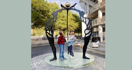 Två kvinnor står inne i ett konstverk som består av gestalter i gjutjärn som håller i en boll