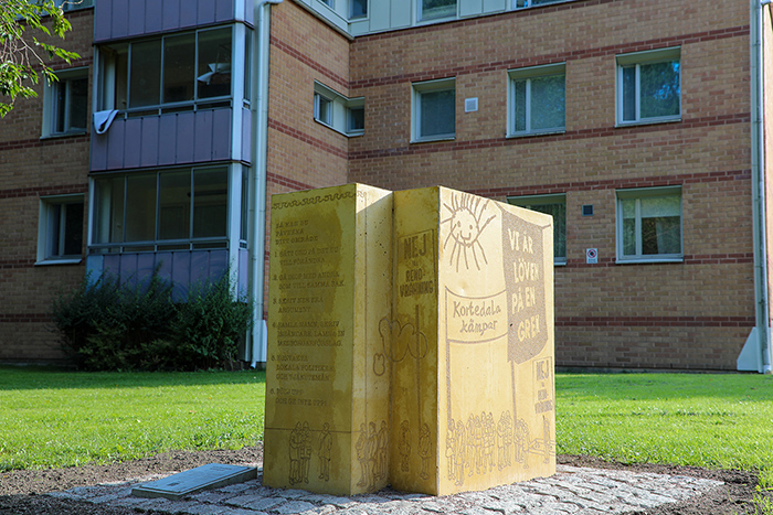 Ett gult monument i betong med bilder av en sol och tv&aring; banderoller som det st&aring;r "Kortedala k&auml;mpar" och "Vi &auml;r l&ouml;ven p&aring; en gren" p&aring;. Ett plakat med texten "NEJ till renovr&auml;kning" syns.