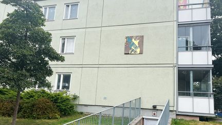 Ett förgyllt glasmotiv i blått och gult hänger på en husfasad i betong.