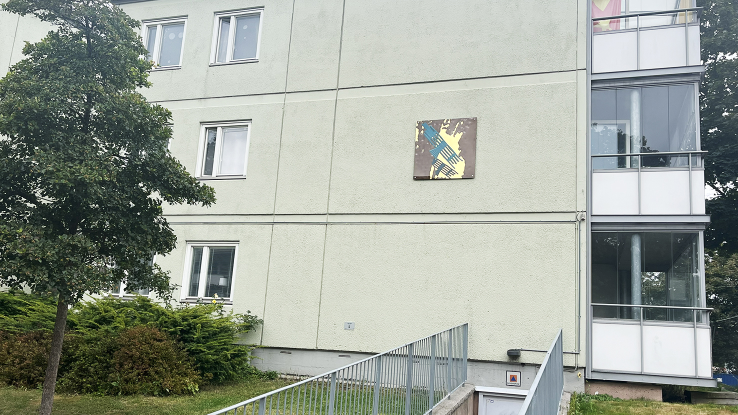 Ett förgyllt glasmotiv i blått och gult hänger på en husfasad i betong.
