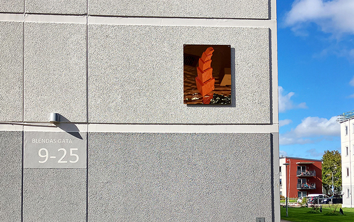 Ett f&ouml;rgyllt glasmotiv som h&auml;nger p&aring; fasaden av ett betonghus. Motivet f&ouml;rest&auml;ller ett r&ouml;tt abstrakt torn.