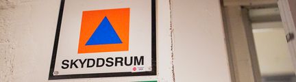 Skylt med texten SKYDDSRUM med en blå triangel i en orange fyrkant.