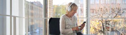 En kvinna sitter i en fåtölj på Stadsbiblioteket. Hon tittar ner i en telefon och har hörlurar på sig.