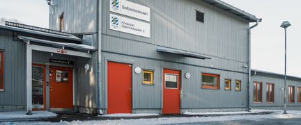 En grå skolbyggnad med röda dörrar och skyltarna Solbackeskolan och Förskolan Stjärnbildsgatan 2 på fasaden.