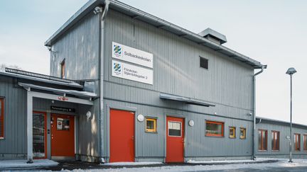 En grå skolbyggnad med röda dörrar och skyltarna Solbackeskolan och Förskolan Stjärnbildsgatan 2 på fasaden.