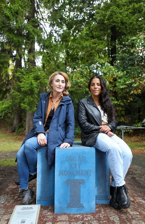 Tv&aring; kvinnor sitter p&aring; en bl&aring; betongkloss med texten "Jag &auml;r ett monument".
