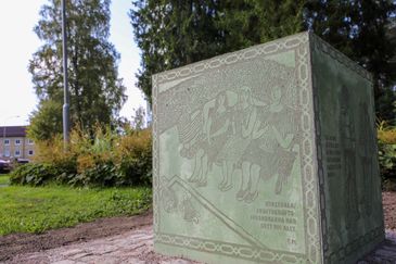 En grön, rektangulär betongkloss med bild och text, framför gräs, buskar och träd.