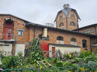 Gamla byggnad i rött tegel med ett torn, framför byggnaden finns gröna odlingar