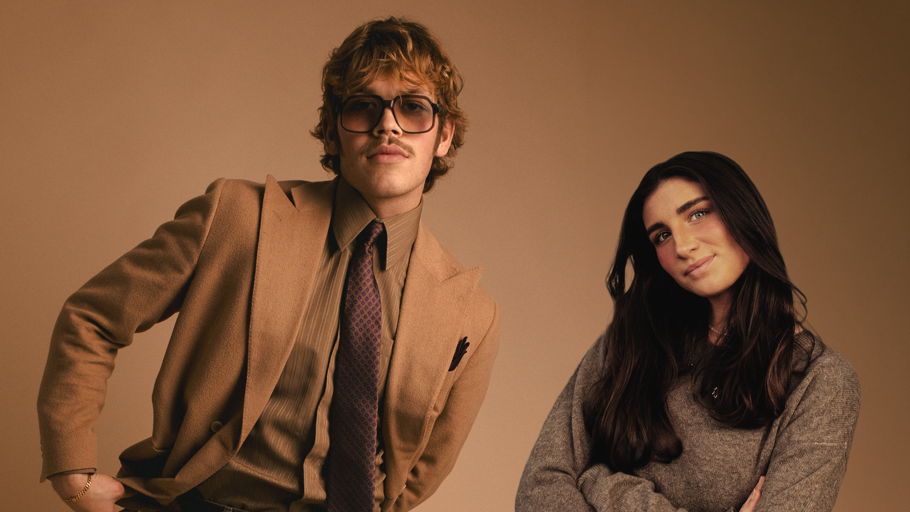 Två unga människor i halvbild, en lockigt blont hår, glasögon, kavaj och slips, en i mörkt långt hår och grå tröja