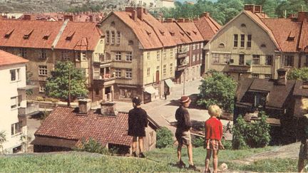 Kolorerat vykort med tre barn i förgrunden. I bakgrunden ett kvarter med landshövdingehus.