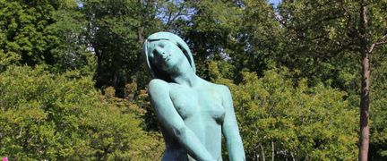 Närbild på övre delen av en skulptur föreställandes en kvinna, med träd och buskar i bakgrunden.