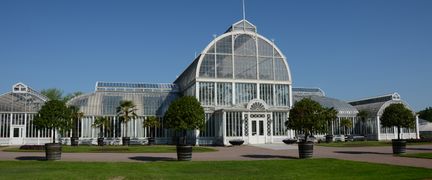 Palmhuset i vitt gjutjärn och glas, med små träd i krukor utanför.