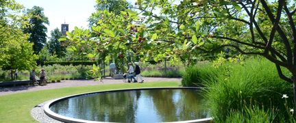 Rund damm mitt på gräsmatta i Trädgårdsföreningen. Stig med personer som promenerar förbi i bakgrunden.