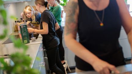 Två barn och en vuxen står vid en spis och lagar mat. En person med svart linne står i längst fram i bilden och skär mat.