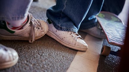 Närbild på två ungdomars vita skor och jeans och en skateboard.