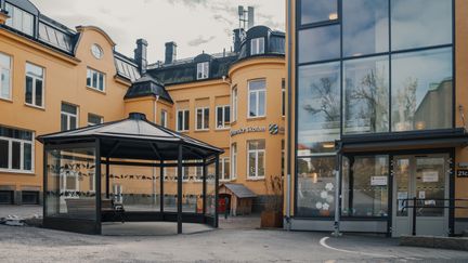 En paviljong med en bänk i framför en gul skolbyggnad med en entré