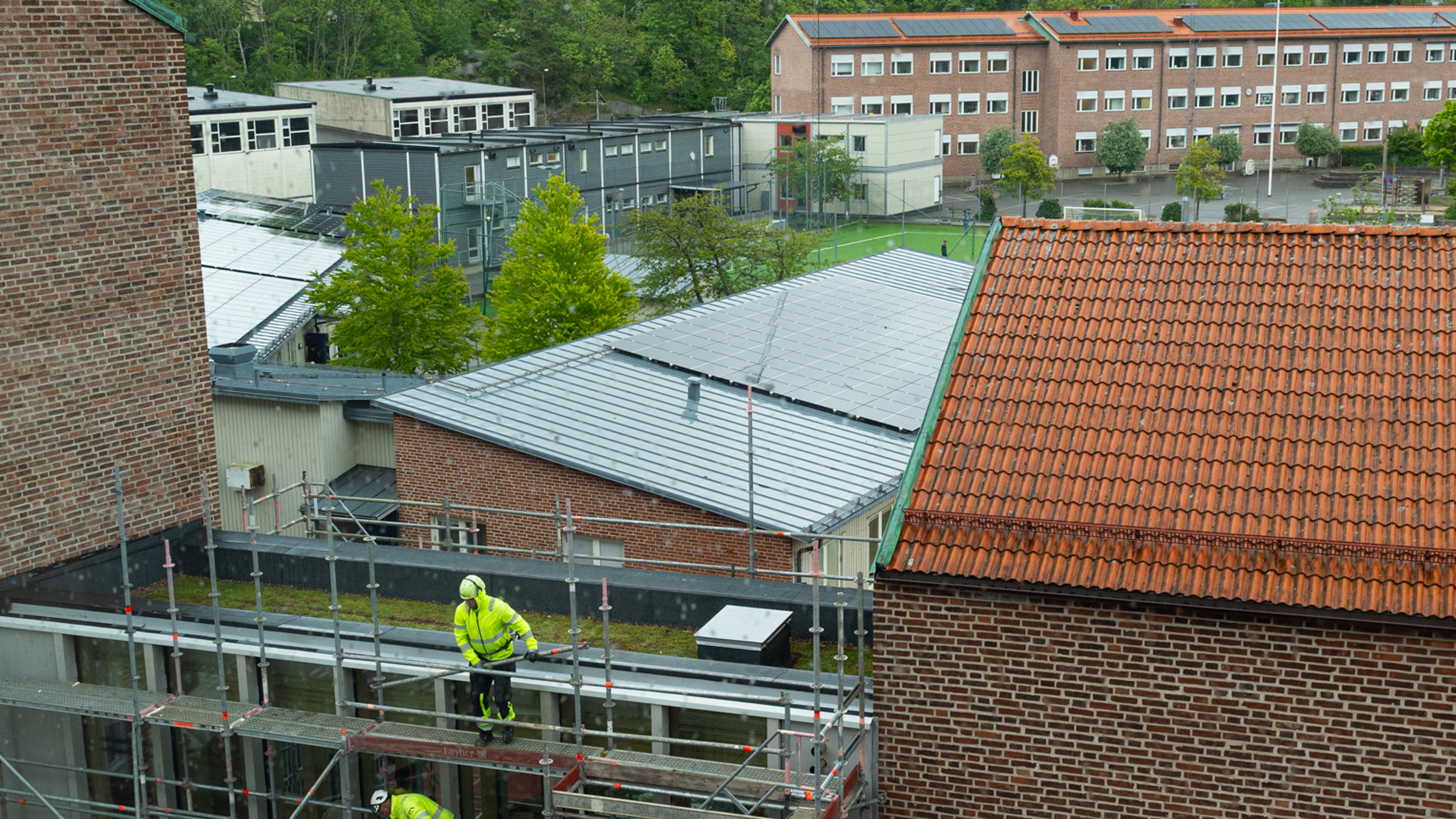 Två personer i varselkläder tar ner en byggställning på en byggnad som binder ihop två andra byggnader. I bakgrunden ser man en skolbyggnad.