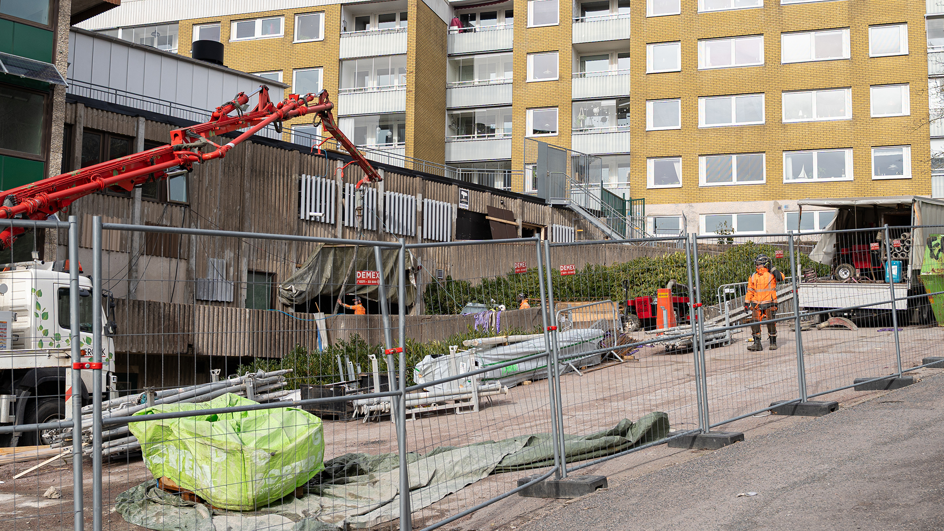 Maskiner och personer i varselkl&auml;der st&aring;r innanf&ouml;r ett byggstaket och arbetar med en betongbyggnad.