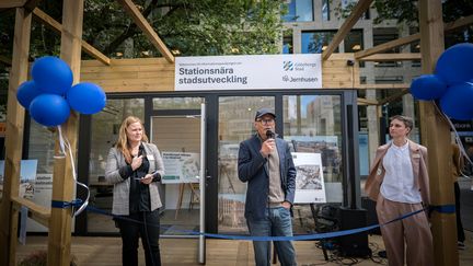 Tre personer står framför en glaspaviljong och håller mikrofoner. Ett blått band som ska klippas är uppspänd framför entrén. På väggen bakom syns en skylt om stadsutveckling.