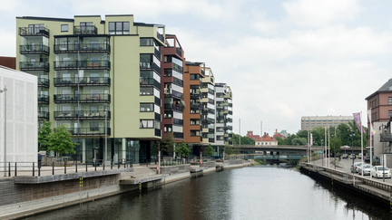 Flera hus i 6 våningar i olika färger, som grönt, brunt och vitt. De ligger längsmed en å. Det finns en mindre gångbro över ån och i fjärran ser man ikonbyggnaden SKFs gamla kullagerfabrik i Gamlestaden.