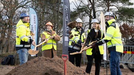Fem personer i varseljackor och bygghjälmar gräver i en sandhög med flerskaftade spadar.