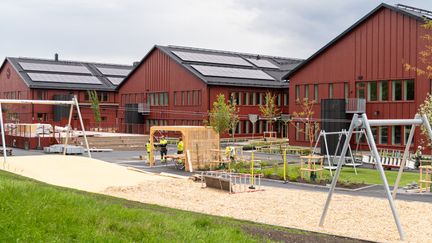 En röd träbyggnad i tre sektioner med solceller på taket. I förgrunden en lekplats.