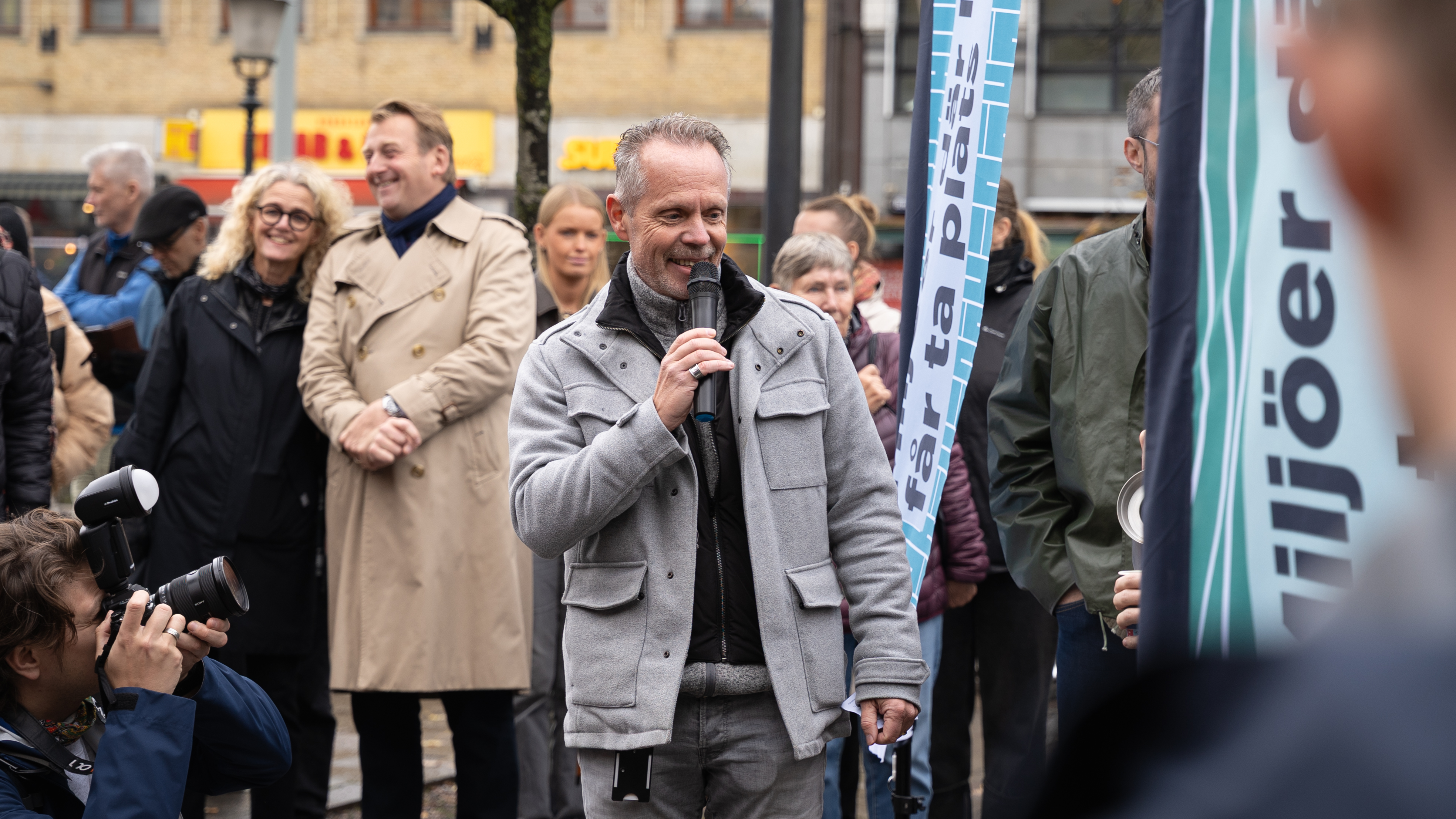 En man som pratar i en mikrofon blir fotograferad av en person som hukar framf&ouml;r honom. I bakgrunden st&aring;r glada m&auml;nniskor.
