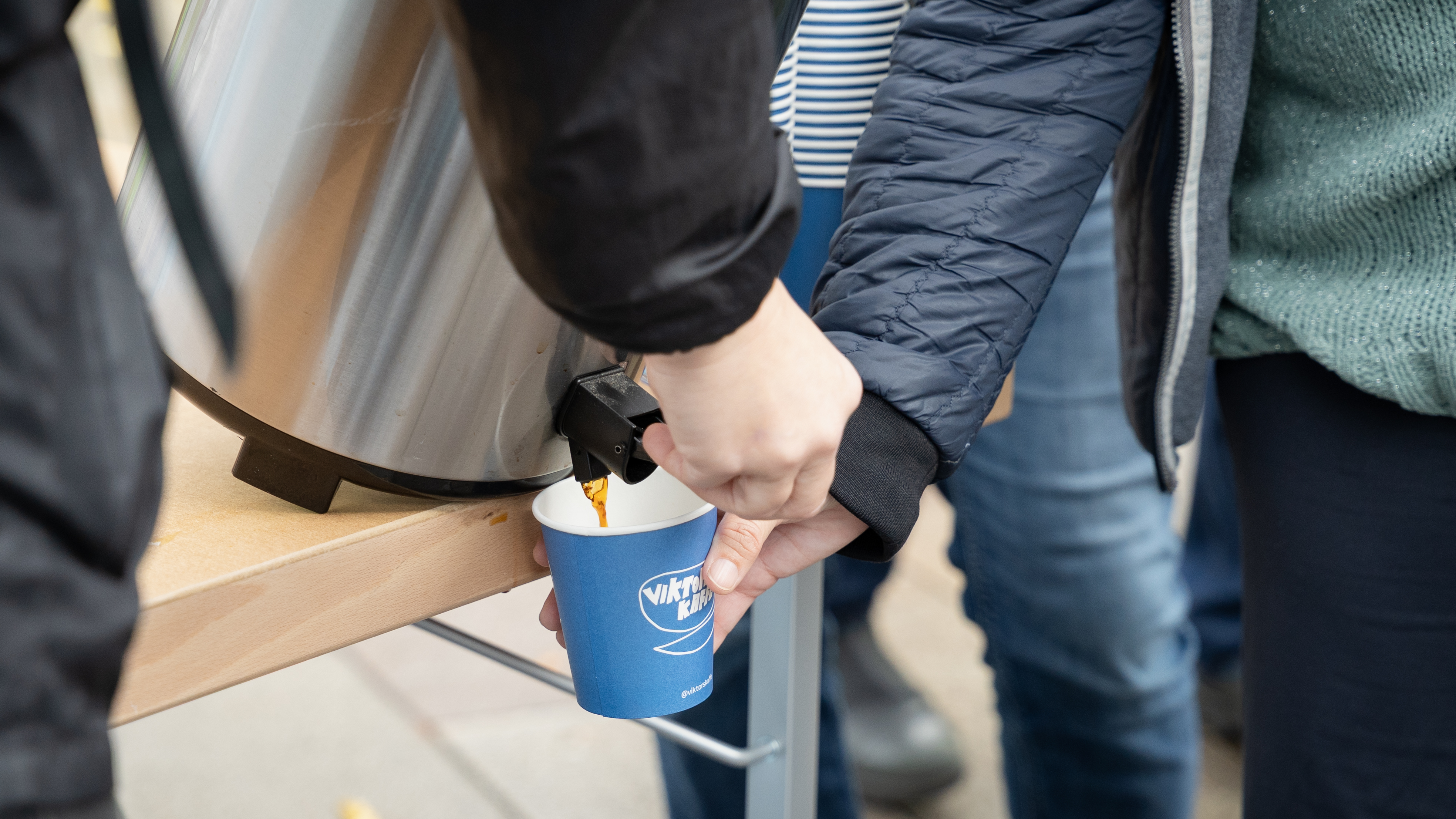 N&aring;gon h&auml;ller upp kaffe i en bl&aring; pappmugg med texten Viktors kaffe, ur en termos som lutas f&ouml;r att f&aring; ut det sista.