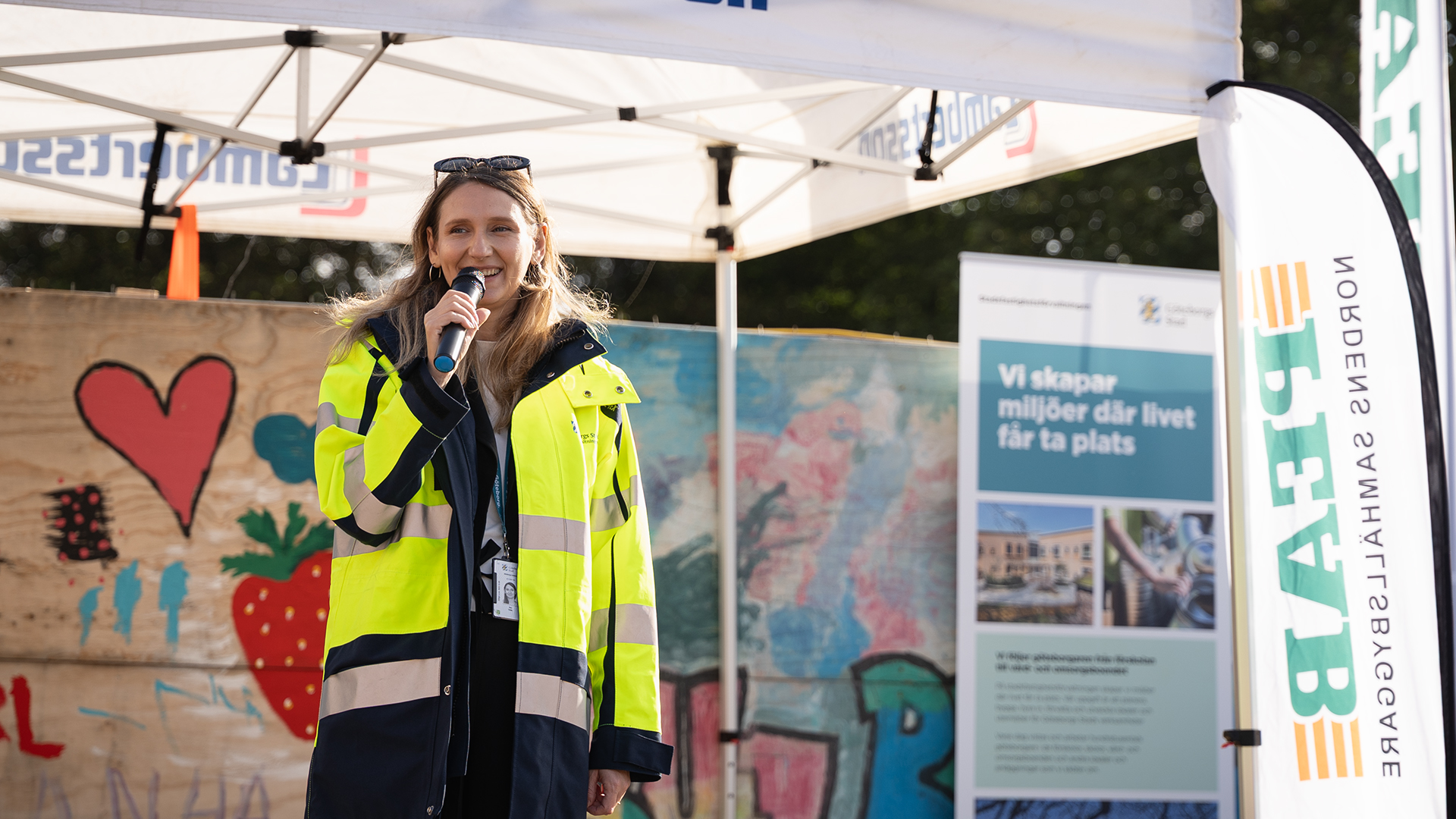 En kvinna i varseljaccka med mikrofon i handen, framför ett byggstaket med målningar på.