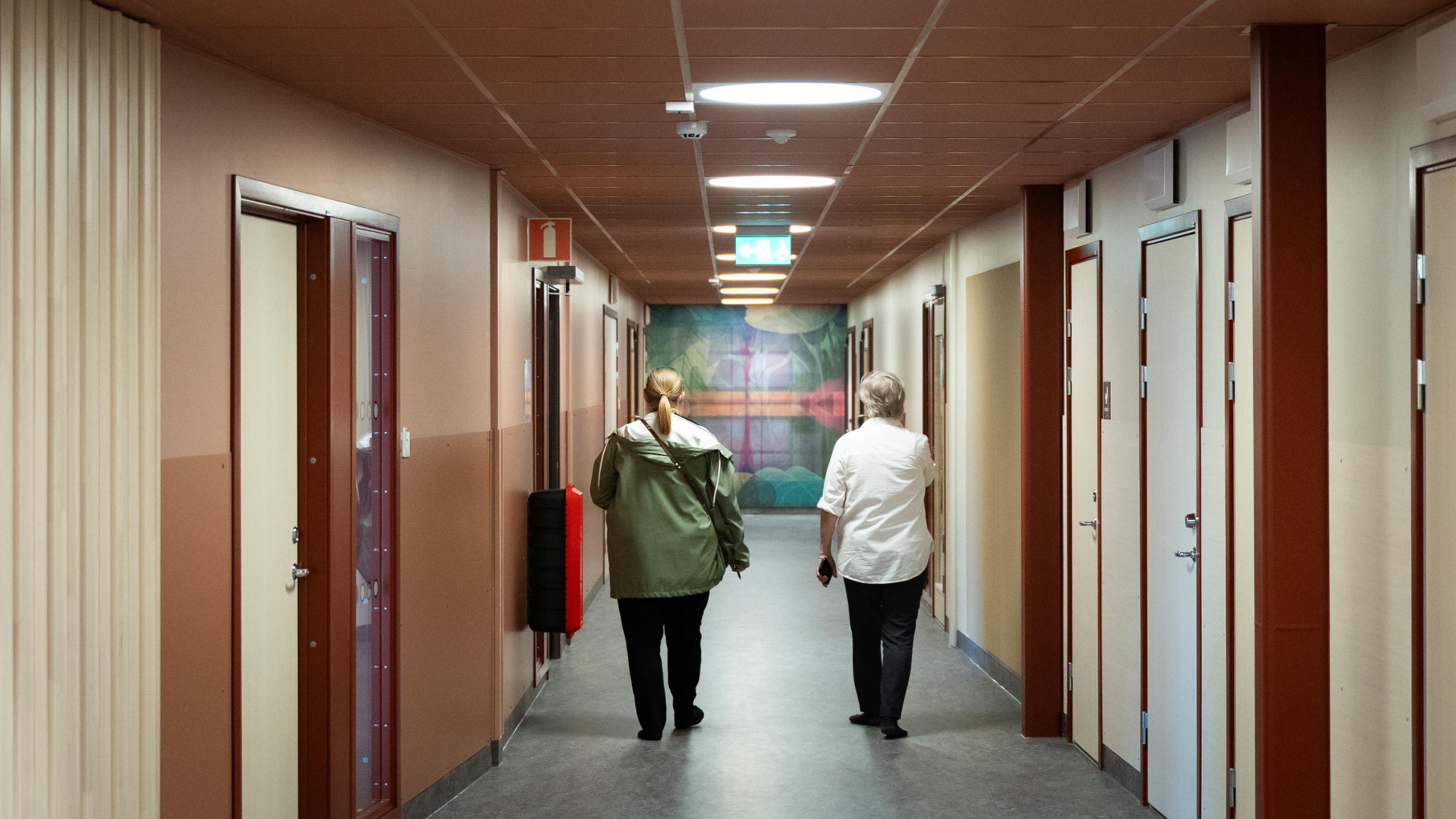 Två personer går med ryggarna mot kameran i en korridor där tak och väggar går i rött och beigegult.