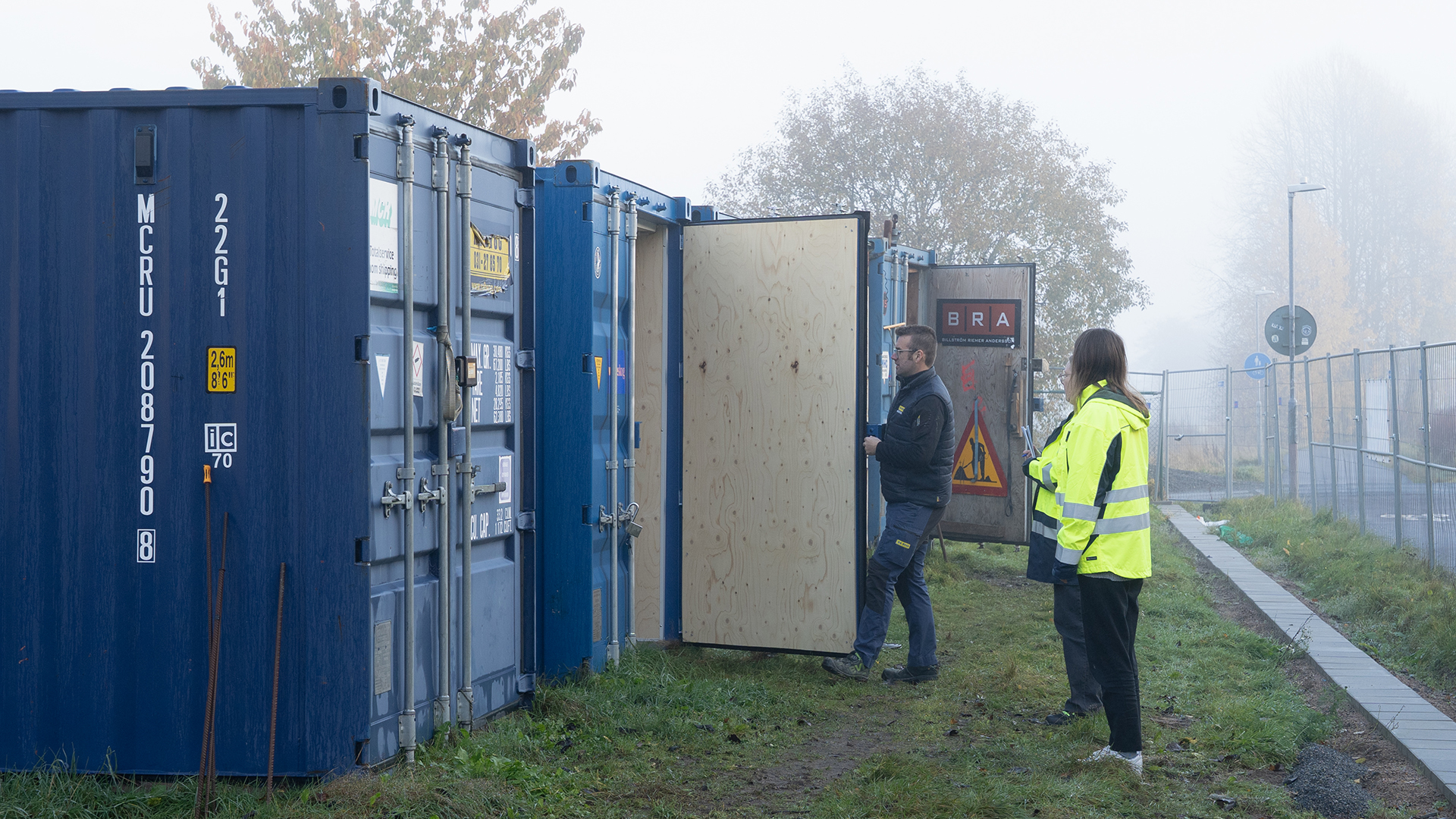 Tv&aring; personer st&aring;r och tittar in i n&aring;gra bl&aring;a fraktkontainrar som har &ouml;ppnna d&ouml;rrar.