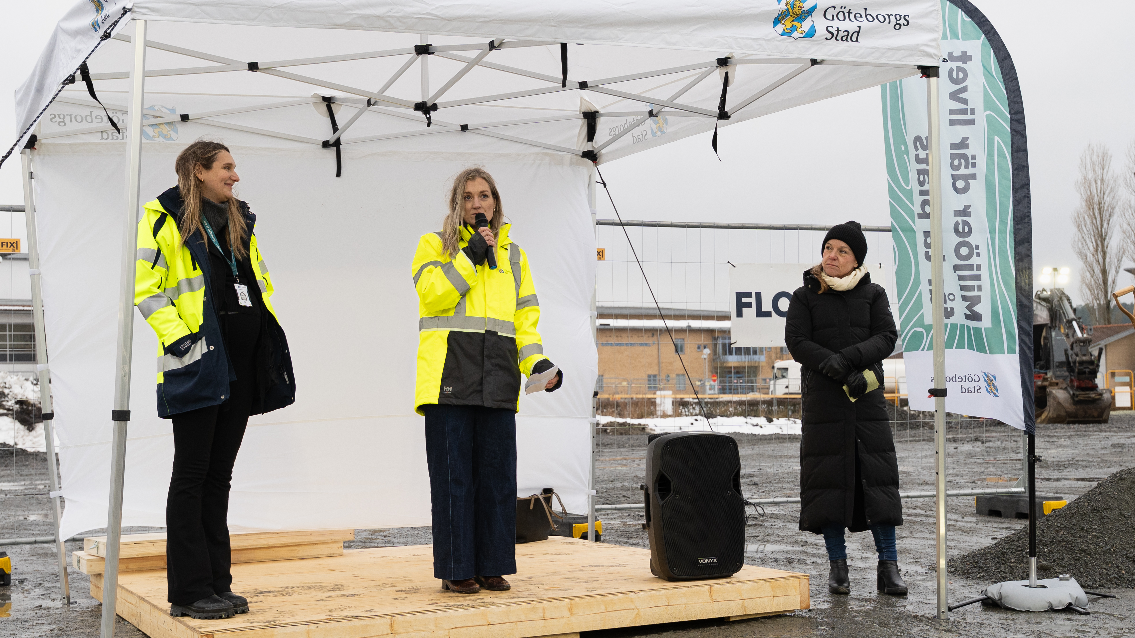 Tv&aring; kvinnor i varseljackor st&aring;r p&aring; en liten scen och pratar i en mikrofon. Bredvid st&aring;r ytterligare en kvinna.. I bakgrunden syns en gr&auml;vmaskin.