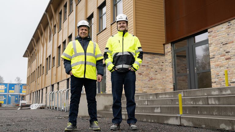 Två personer i varselkläder och bygghjälmar står utanför en gul trevåningsbyggnad.